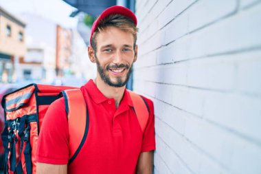 Beyaz teslimatçı, kırmızı üniforma giyiyor ve sırt çantası teslim ederken gülümsüyor.
