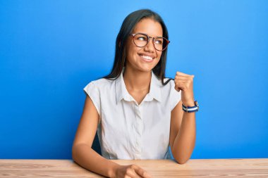 Güzel İspanyol kadın, gündelik giysiler giyip masaya oturmuş gülümsüyor, gülümsüyor ve başparmağıyla yan tarafa bakıyor.. 