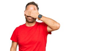 Yakışıklı, orta yaşlı, sıradan kırmızı tişörtler giyen, gülümseyen ve elleri yüzünde, şaşırtmak için gözleri kapatan bir adam. kör kavram. 