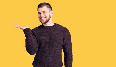 Gündelik süveter giyen, neşeli bir şekilde gülümseyen, avuç içiyle kameraya bakan genç yakışıklı bir adam.. 