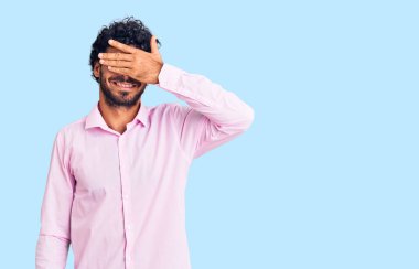 Kıvırcık saçlı ve ayı gibi iş kıyafetleri giyen yakışıklı bir genç adam. Gülümsüyor ve elleri yüzünde, şaşırtmak için gözlerini kapatıyor. kör kavram. 