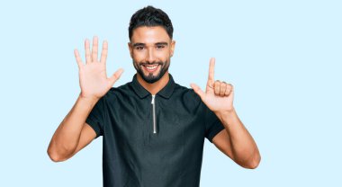 Sakallı genç adam spor giyer ve yedinci parmaklarını göstererek mutlu ve kendinden emin bir şekilde gülümser.. 