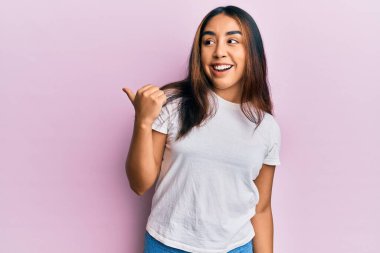 Gündelik beyaz t-shirt giyen genç Latin kadın başparmağını yana kaldırıyor ve ağzı açık gülümsüyor. 