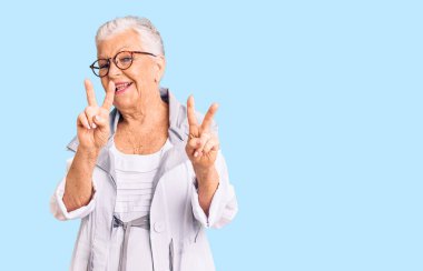 Mavi gözlü, gri saçlı, sıradan kıyafetler giyen, gülümseyen ve parmaklarını zafer işareti yaparken gösteren kameraya bakan son sınıf güzeli bir kadın. İki numara.. 