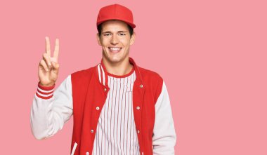 Beyzbol üniforması giyen yakışıklı beyaz adam iki numarayı gösterip işaret ederken kendinden emin ve mutlu bir şekilde gülümsüyor.. 