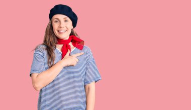 Genç, sarışın, Fransız beresi takan, çizgili, neşeli tişörtlü, yüzünde bir gülümseme olan, mutlu ve doğal bir ifadeyle elini ve parmağını kaldıran genç bir kadın. 