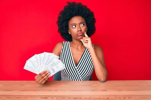 Young african american woman holding dollars serious face thinking about question with hand on chin, thoughtful about confusing idea