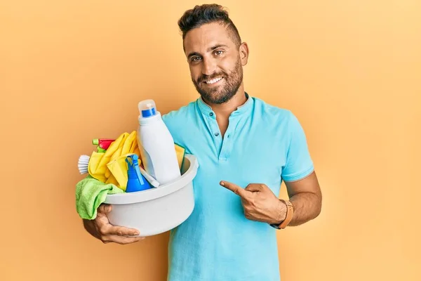 Handsome man with beard holding cleaning products smiling happy pointing with hand and finger