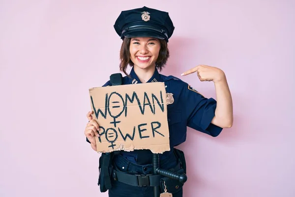 Young beautiful girl wearing police uniform holding woman power banner pointing finger to one self smiling happy and proud