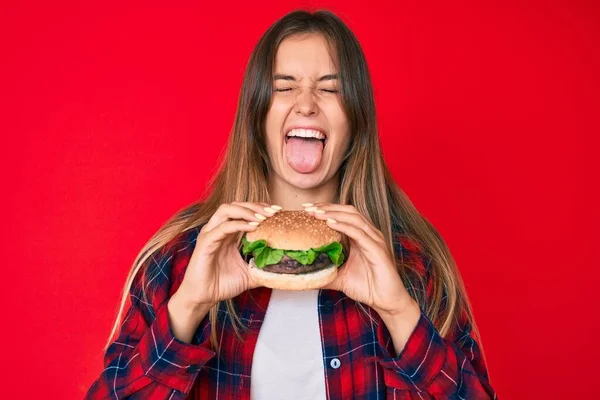 美しいですCaucasian女性食べるおいしい古典的なハンバーガー突き出た舌で幸せな面白い式 — ストック写真