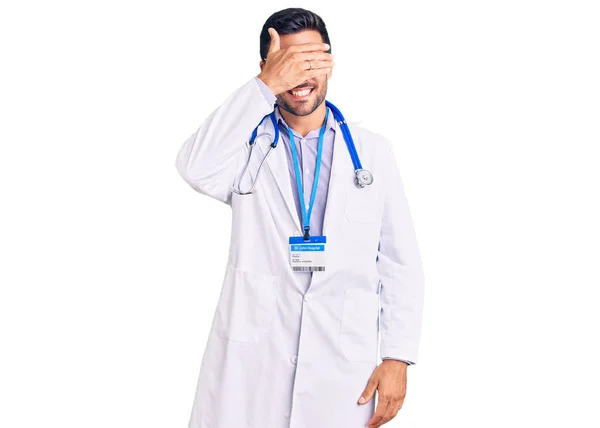 医者の制服を着た若いヒスパニック系の男が笑顔と驚きのために目をカバー顔に手で笑っています 視覚障害の概念 — ストック写真