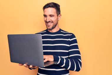 Genç yakışıklı adam dizüstü bilgisayarını kullanarak sarı arka planda duruyor. Pozitif ve mutlu görünüyor. Kendine güvenen bir gülümsemeyle dişlerini gösteriyor.