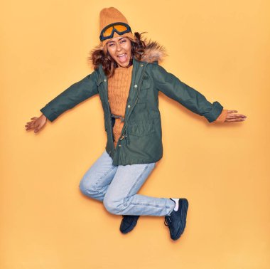 Kışlık giysiler giyen genç, güzel Latin kadın mutlu gülümsüyor. Yüzünde ve kollarında tebessümle, izole edilmiş sarı arka planda atlıyor.