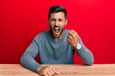 Yakışıklı, gündelik tarzda giyinmiş, kızgın ve kızgın bir şekilde masaya oturmuş öfkeyle bağırırken yumruğunu sallıyor. öfke ve agresif konsept. 
