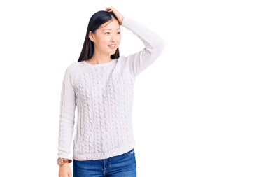 Genç, güzel, Çinli bir kadın, rahat bir süveter giyiyor, gülümsüyor, saçlarına dokunuyor, el hareketi yapıyor, çekici ve modaya uygun poz veriyor. 