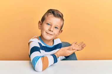 Gündelik giysiler giyen sevimli beyaz çocuk masaya oturmuş gülümseyerek içeri girmeye davet ediyor. 