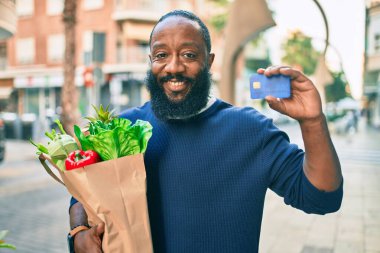 Sakallı Afrikalı Amerikalı bir adam elinde süpermarketten alınmış kağıt torbalarla kredi kartı gösteriyor.