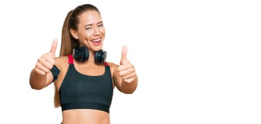 Güzel sarışın kadın spor kıyafetleri giyiyor ve kulaklık takıyor. Elleriyle olumlu jest yapıyor. Gülümsüyor ve başarı için mutlu. kazanan jest. 