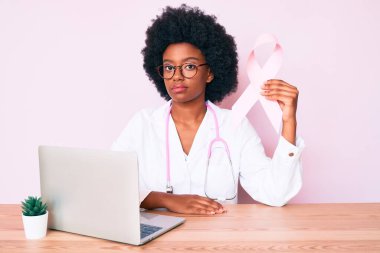 Genç Afrikalı Amerikalı kadın doktor üniforması giyiyor. Elinde pembe kanser kurdelesi var. Düşünme tavrı takınıyor. 
