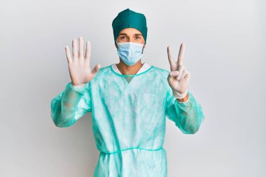 Genç, yakışıklı, cerrah üniforması giyen ve sağlık maskesi takan yedi numaralı parmaklarıyla gülümseyerek mutlu ve kendinden emin.. 