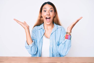 Genç esmer kadın gündelik giysiler içinde masada oturuyor zaferi kutluyor mutlu bir gülümseme ve ellerini kaldırarak kazanan yüz ifadesi. 