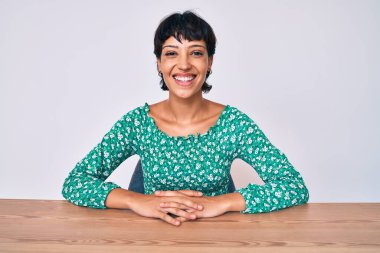 Güzel esmer kadın, sıradan kıyafetler giyip mutlu ve havalı bir gülümsemeyle masada oturuyor. şanslı kişi. 