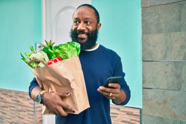 Sakallı Afrikalı Amerikalı adam elinde süpermarketten alınmış kağıt torbalarla akıllı telefon kullanıyor.