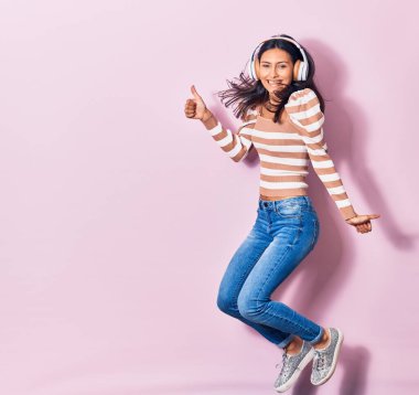 Mutlu gülümseyen genç, güzel İspanyol kadın. Yüzünde tebessümle zıplıyor kulaklık takıyor pembe arka planda davullarla imza atıyor.