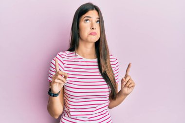 Genç esmer kadın pembe arka plan üzerine günlük giysiler giymiş üzgün ve üzgün görünüyor, parmaklarıyla yön gösteriyor, mutsuz ve depresif.. 