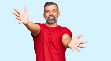 Orta yaşlı, sıradan kıyafetler giyen yakışıklı bir adam kameraya bakıp sarılmak için kollarını açmış gülümsüyor. Mutluluğu kucaklayan neşeli bir ifade.. 