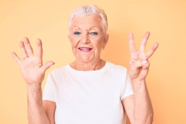 Son sınıf güzeli, mavi gözlü, gri saçlı, sarı arka planda klasik beyaz tişört giyen, 8 numaralı parmaklarıyla işaret eden, kendinden emin ve mutlu gülümseyen bir kadın.. 