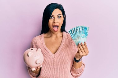 Kafkasyalı genç bir kadın Brezilya banknotları ve domuz kumbarası tutuyor. Şaşırmış ve şaşırmış bir ifade, korku ve heyecanlı bir yüz ifadesiyle.. 