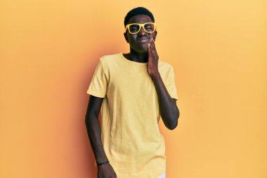 Sıradan kıyafetler giyen ve gözlüklü genç Afro-Amerikan bir adam diş ağrısı ya da diş hastalığından dolayı ağızlarına acı dolu bir ifadeyle dokunuyor. dişçi 