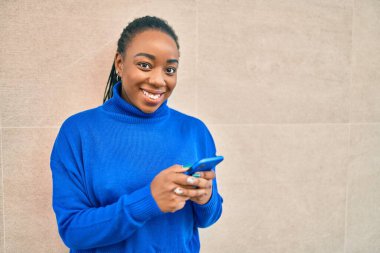 Genç Afrikalı Amerikalı kadın şehirde akıllı telefon kullanarak mutlu bir şekilde gülümsüyor..