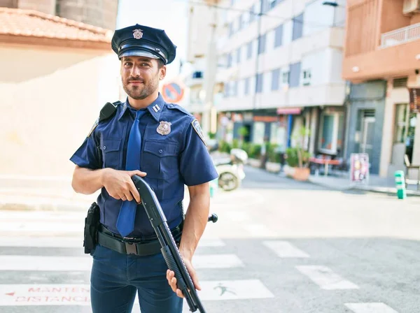 年轻英俊的惊慌失措的警察穿着警服开心地笑着 站在街上 面带微笑地拿着猎枪 — 图库照片