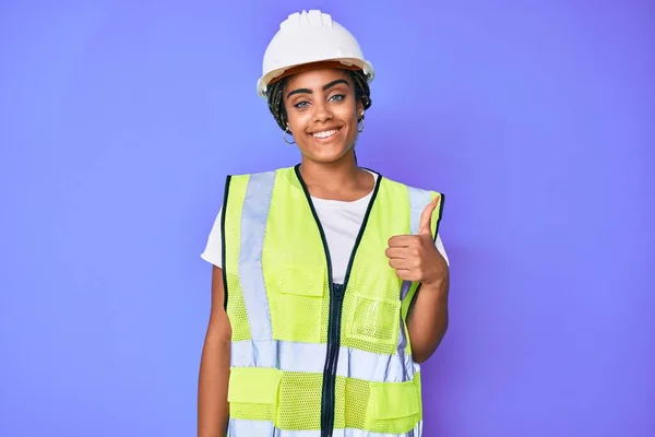 Young african american woman with braids wearing safety helmet and reflective jacket smiling happy and positive, thumb up doing excellent and approval sign