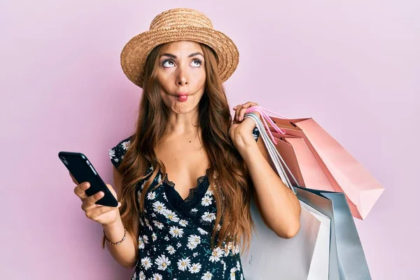 Young brunette woman holding shopping bags and smartphone making fish face with mouth and squinting eyes, crazy and comical.