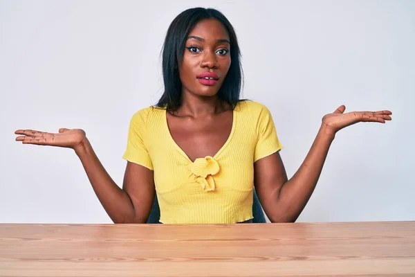 身穿休闲装的年轻非洲裔美国女人坐在桌子上 毫无头绪 与张开的双臂混淆不清 毫无头绪 满脸疑惑 — 图库照片