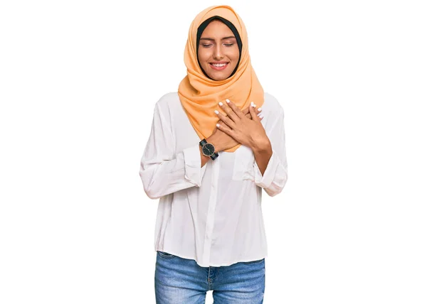 伝統的なイスラム教のヒジャーブスカーフを身に着けている若いブルネットの女性は 目を閉じて胸に手で笑顔と顔に感謝のジェスチャー 健康の概念 — ストック写真