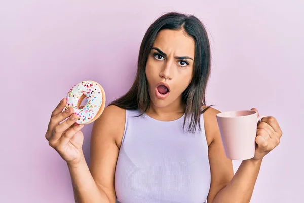 年轻的黑发女人吃着甜甜圈 脸上带着震惊的表情喝咖啡 脸上带着怀疑和讽刺的表情 张开嘴让人吃惊 — 图库照片