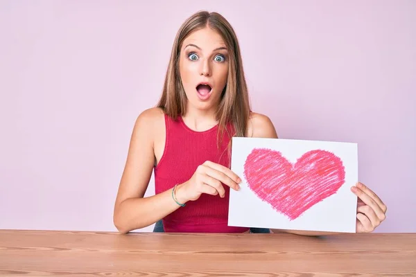 Joven Chica Rubia Sentada Mesa Sosteniendo Corazón Dibujar Miedo Sorprendido —  Fotos de Stock