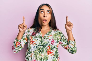 Gündelik kıyafetler giyen genç Latin kadın şaşırmış ve şaşırmış bir şekilde yukarı bakıyor ve parmaklarıyla işaret ediyor ve kollarını kaldırıyordu.. 