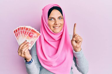Kafkasyalı genç bir kadın, elinde 20 şekel banknot tutan İslami başörtüsü takıyor. Bir fikir veya soruyla gülümsüyor. 