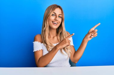 Güzel sarışın genç kadın, gündelik giysiler içinde masada oturuyor gülümsüyor ve kameraya bakıyor. İki eliyle ve parmaklarıyla kenarı işaret ediyor.. 