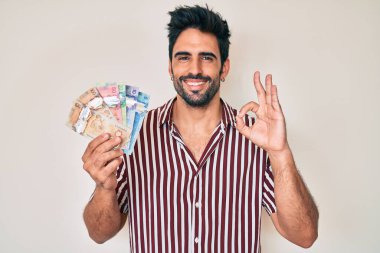 Sakallı yakışıklı İspanyol adam Kanada doları tutuyor. Parmaklarıyla imza atıyor, gülümsüyor. 
