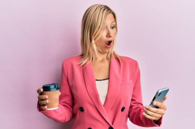 Güzel orta yaşlı sarışın kadın akıllı telefon kullanıyor ve şok olmuş bir şekilde bir fincan kahve içiyor. Şüpheci ve alaycı görünüyor. 