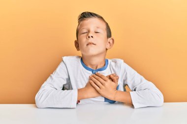 Masada oturan, elleri göğsünde, gözleri kapalı ve yüzünde minnet dolu bir jestle gülümseyen beyaz sevimli bir çocuk. sağlık kavramı. 