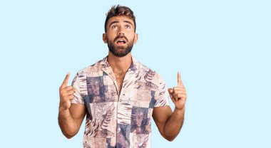 Yaz kıyafetleri giyen genç İspanyol adam şaşırmış ve şaşırmış bir şekilde yukarı bakıyor ve parmaklarıyla işaret ediyor ve kollarını kaldırıyordu.. 