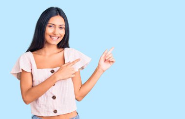 Gündelik kıyafetler giyen, gülümseyen ve kameraya bakan genç, güzel bir Latin kız. İki elini ve parmağını kenarı işaret ediyor.. 