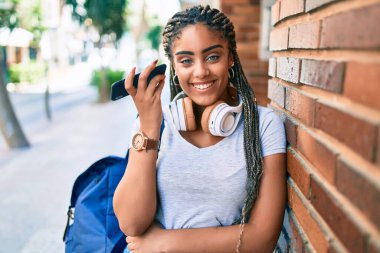 Genç Afrikalı Amerikalı öğrenci gülümseyerek üniversite kampüsünde sesli mesaj dinliyor.